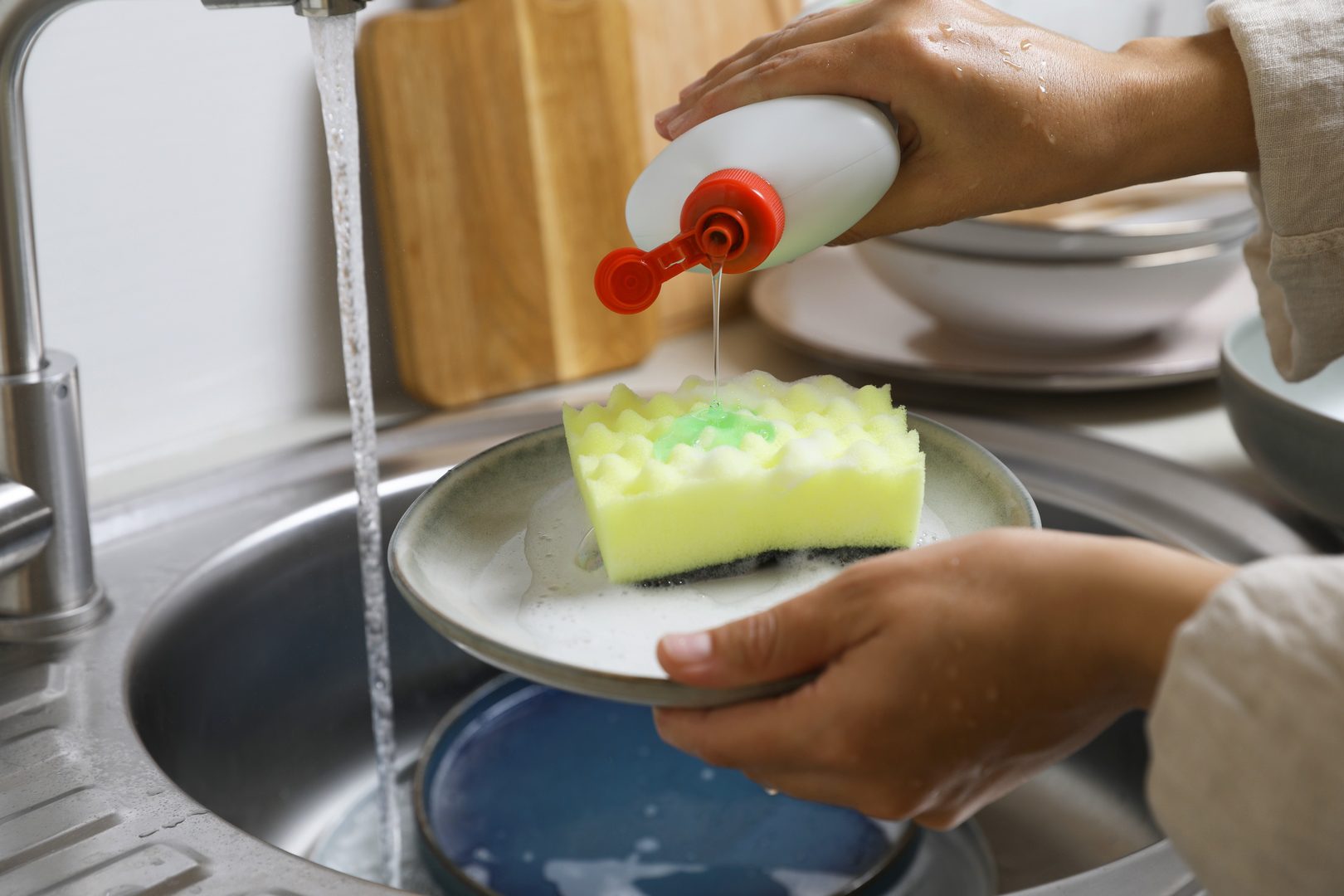 Woman pouring dishwashing detergent onto plate with sponge near kitchen sink, closeup